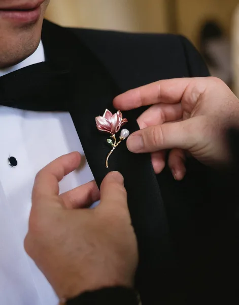 Groom boutonniere being pinned, a pink boutonniere with pearl accent and gold stem pin on a black tuxedo in warm indoor light