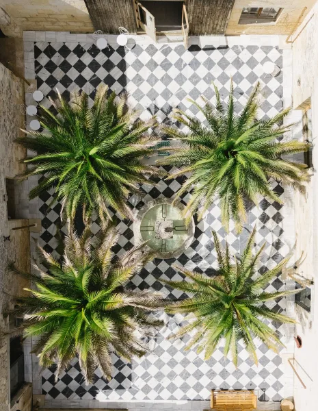 Venue courtyard overhead view of a checkerboard tile floor with palm trees and a fountain, surrounded by café tables, chairs, and stone walls