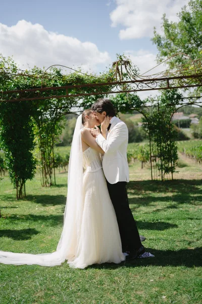 Wedding kiss portrait of bride and groom kissing beneath a vine-covered arbor, bride’s long veil flowing against rolling hills and sky