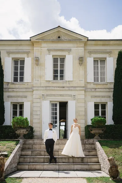 Couple portrait of bride in strapless gown and groom in white tuxedo with bow tie posing on stone estate steps under blue sky