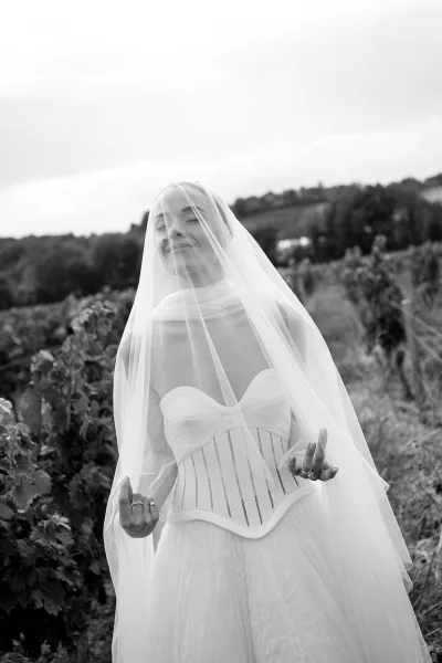 Bridal portrait of a bride with veil over face, eyes closed, in a strapless corset dress among vineyard rows and hillsides under sky
