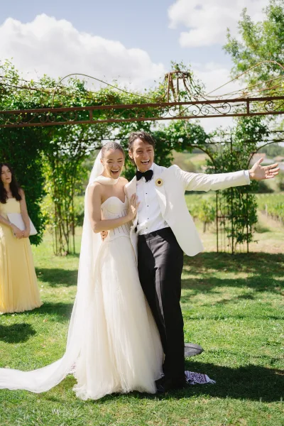 Couple portrait of bride and groom laughing under a vine-covered garden arbor, her long veil flowing as he extends his arm in a tuxedo