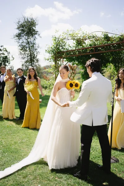 Ceremony moment as bride holds sunflowers and the groom’s hand beneath a metal garden arbor, wedding party lined up behind them on the lawn