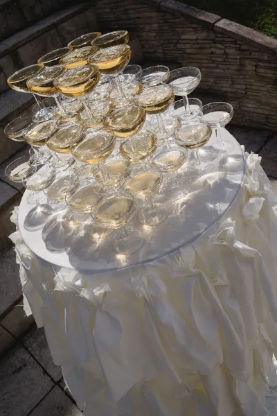 Champagne tower of coupe glasses stacked on a round table with white tablecloth and fabric rosettes on an outdoor patio stone wall