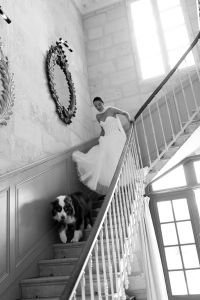 Bridal portrait of a bride walking downstairs in a strapless corset tulle gown, scarf wrap flowing, with a dog in the hallway by stone stairs