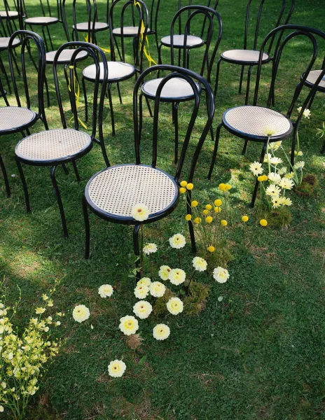 Ceremony seating with black bentwood chairs and cane seats tied with yellow ribbon beside wildflower ground arrangements on a garden lawn