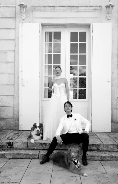 Couple portrait in a black and white wedding portrait style, bride in strapless gown beside groom on stone steps with their dogs by French doors
