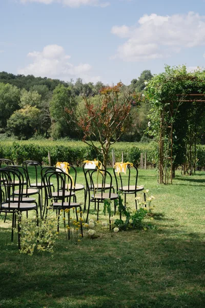 Ceremony setup for an outdoor wedding ceremony with black chairs and yellow ribbon bows facing a floral arch on a vineyard lawn under blue skies