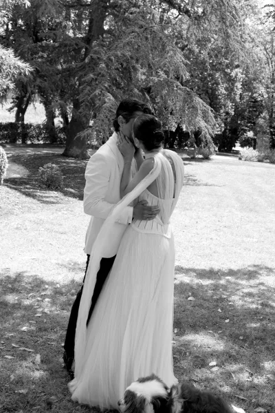 Wedding kiss portrait of bride and groom kissing, her long veil flowing in dappled garden light with their dog on the lawn