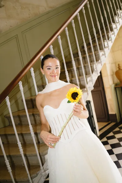 Bridal portrait of a bride holding bouquet of sunflowers, leaning on a staircase banister in a hallway with checkered tile floor