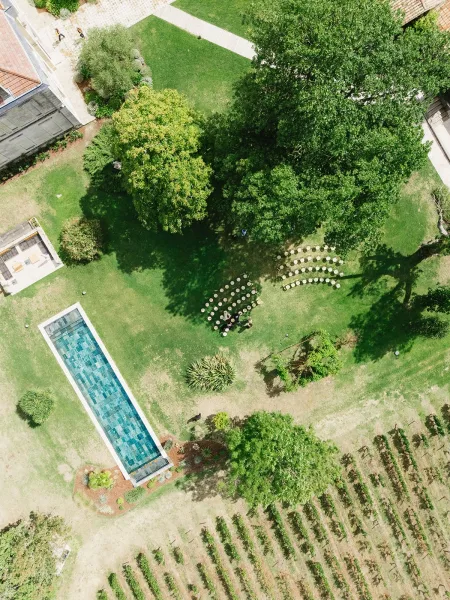 Outdoor ceremony setup with ceremony seating layout, curved rows of chairs on a lawn under trees near a pool and vineyard buildings