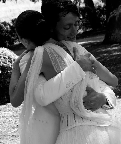 Wedding hug as bride and groom embrace tightly, her strapless gown and ring visible, in sunlit garden trees and lawn background