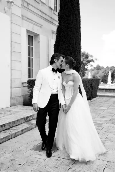 Wedding kiss portrait of bride and groom kiss holding hands, her strapless tulle gown and his white tuxedo on a stone terrace with cypress tree