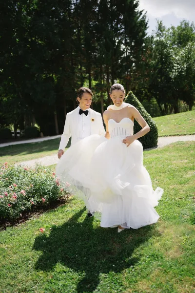 Wedding couple portrait of bride lifting her tulle skirt while walking with groom in a white tuxedo along a sunlit garden path