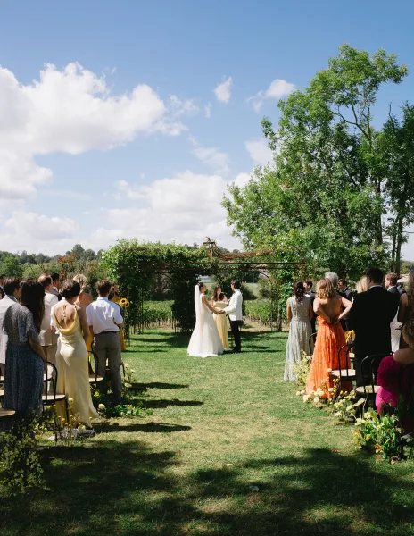 Wedding ceremony with couple exchanging vows under a greenery arch as guests stand on a lawn beside vineyard rows under a blue sky