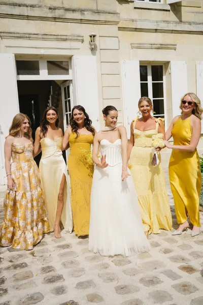 Bride with bridesmaids in yellow dresses posing with bouquets and sunglasses on cobblestone courtyard by stone building doors