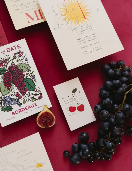 Invitation suite flat lay with illustrated stationery cards and calligraphy text, styled with grapes and fig on burgundy fabric backdrop