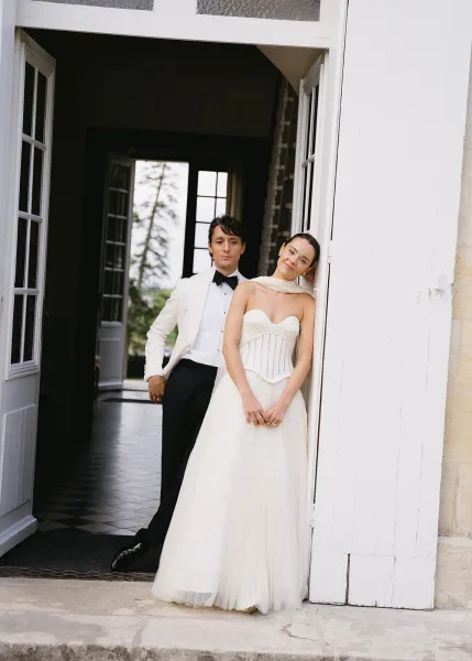 Couple portrait of bride in a corset wedding dress and groom in a white dinner jacket by an open doorway, stone wall and soft window light