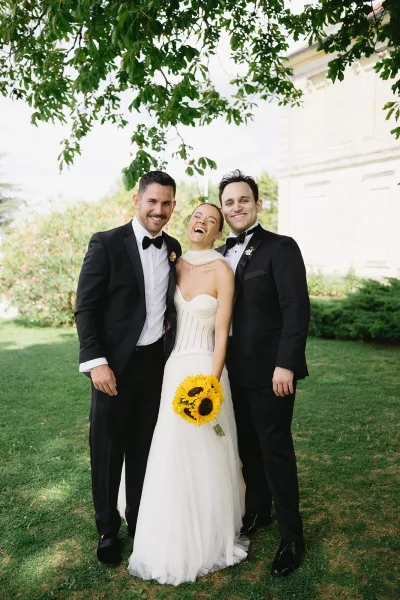 Wedding party portrait of a bride in a strapless gown between two groomsmen in black tuxedos, holding a sunflower bouquet on a green lawn