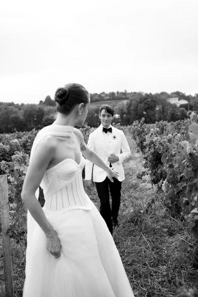 Couple portrait of bride in strapless dress and veil reaching for groom in white tuxedo jacket on a grassy path between vineyard rows