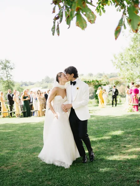 Wedding kiss portrait of bride and groom kissing, her veil flowing beside ceremony chairs on a grassy lawn as guests watch under trees