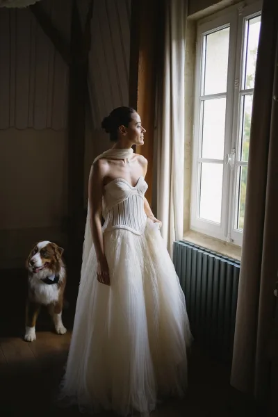 Bridal portrait of a bride by window in a strapless pleated corset bodice and tulle skirt, her dog in a bow tie beside her
