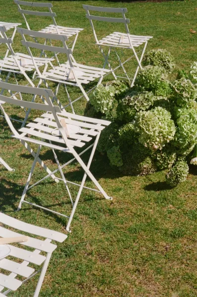 Ceremony seating on a grass lawn features white bistro chairs flanking grounded hydrangeas, framed by simple garden greenery.