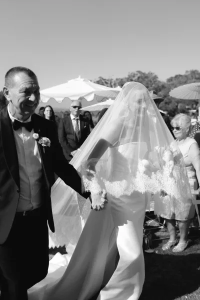Wedding processional with bride walking down aisle on her father's arm, guests shaded by parasols on a sunny lawn beneath open sky and trees.