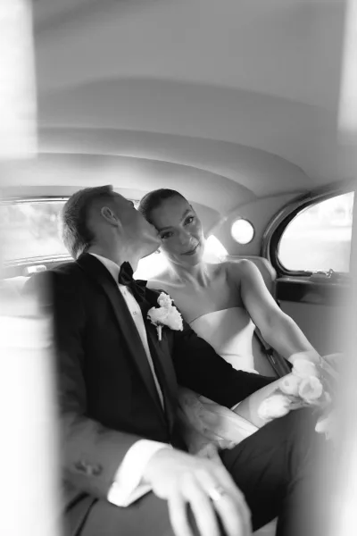 Couple portrait of a bride and groom in a car, framed by a vintage car interior, the bride holding a rose bouquet as light filters through the window.