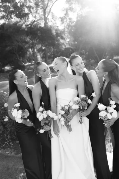 Bridal party portrait of bride and bridesmaids in a garden, holding hand-tied bouquets, with trees and flower bushes along the lawn.