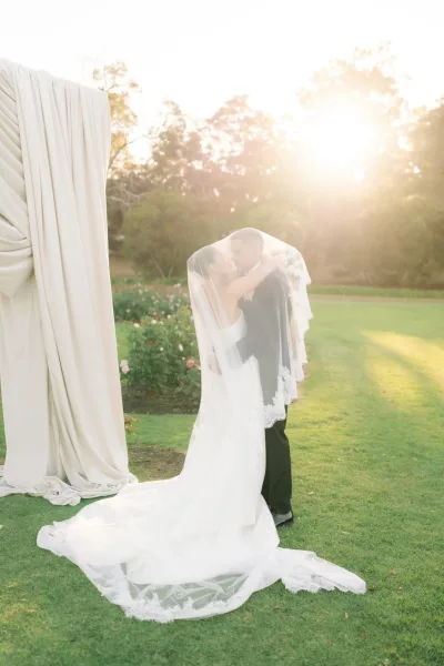 Wedding kiss portrait of newlyweds under the veil, her white bouquet framed by sunlight as they stand on a rose-lined lawn with trees behind.