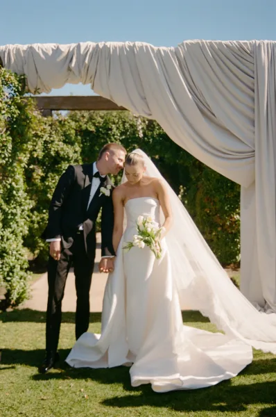 Couple portrait of a bride and groom holding hands beneath a draped fabric arch by a garden pergola.
