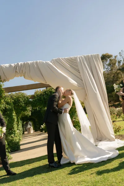 Ceremony kiss during an outdoor ceremony moment, the bride's long veil trailing as they embrace beneath a garden pergola amid hedges and blue sky.