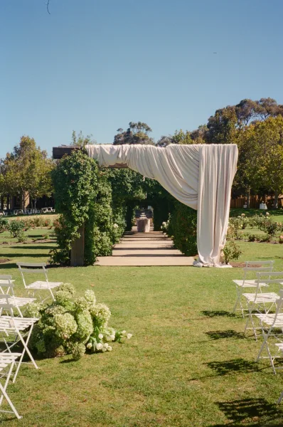 Ceremony setup with outdoor ceremony decor, rows of white chairs on a grass lawn flanked by garden trees and rose bushes under blue sky.