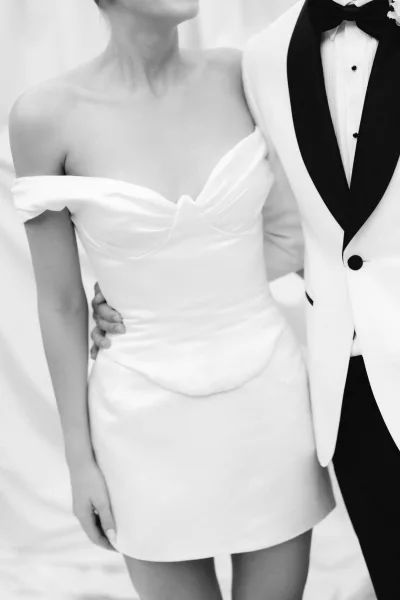 Short wedding dress with an off-the-shoulder bridal neckline and structured bodice, styled with a tuxedo jacket for a bold, modern look.