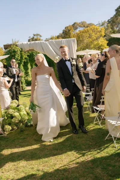 Ceremony exit during the newlyweds’ recessional, the couple holding hands as guests cheer under parasols on a sunlit garden lawn.