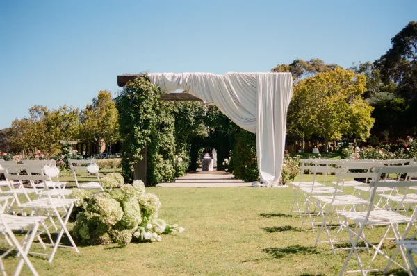 Ceremony setup with outdoor ceremony decor beneath a wooden pergola on a manicured lawn, framed by garden trees under a blue sky.