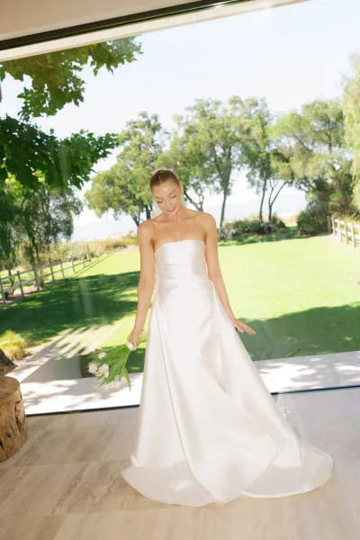 Bridal portrait of a bride holding bouquet, cradling white tulips by a large window, garden trees beyond and stone floor underfoot.