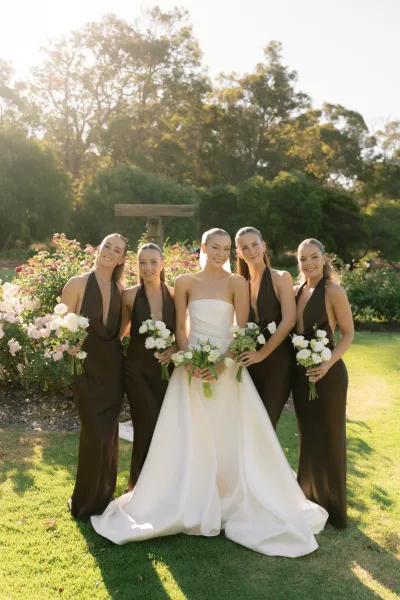 Bride and bridesmaids hold white and green bouquets beneath a wooden arbor amid garden roses, with trees and lawn in the background.