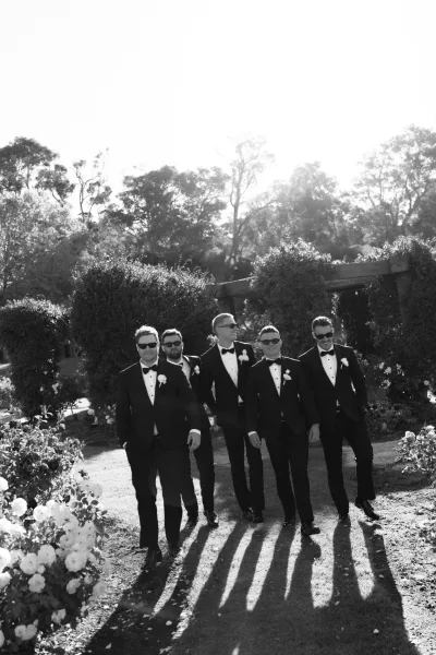 Groomsmen portrait captured as a groomsmen walking photo, sunglasses on as they stroll a rose-lined garden path beneath a pergola and trees.
