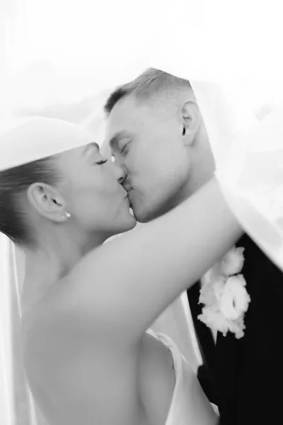 Wedding kiss portrait capturing an under the veil kiss, bride embracing groom with a boutonniere, framed by sheer veil fabric.