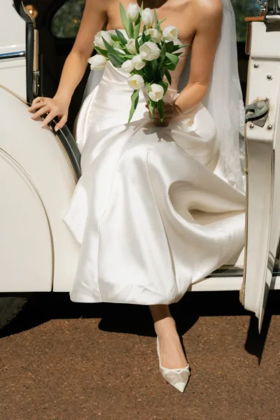 Bridal portrait of a bride with bouquet, cradling white tulips inside a car interior, soft light framing her serene arrival moment.