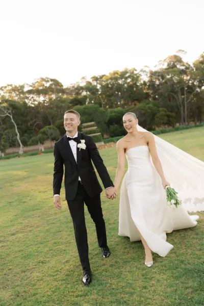 Couple portrait of the bride and groom walking hand in hand outdoors, her veil trailing softly as they share a candid moment.