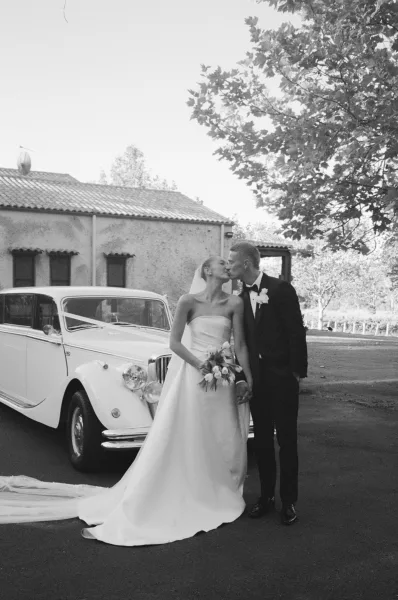 Wedding kiss portrait of a bride and groom with a classic car parked behind them, a timeless, romantic moment framed by soft daylight.