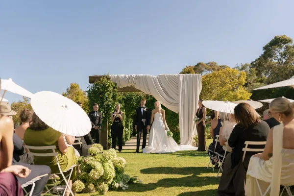 Ceremony moment at an outdoor wedding ceremony as the couple holds hands beneath a fabric-draped arch on a sunlit garden lawn.