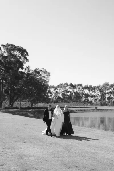 Wedding processional walking down the aisle, with a blusher-veiled bride escorted by her parents across a lakeside lawn under open sky.