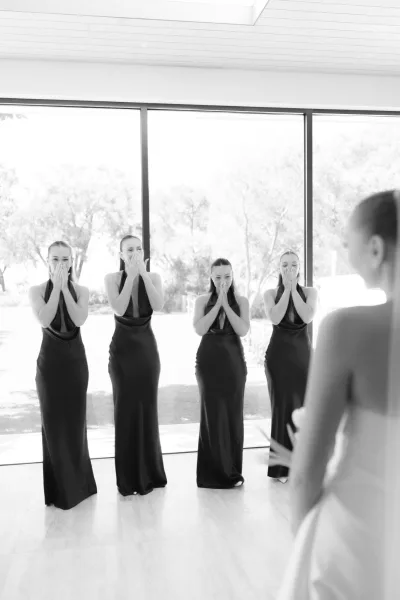 Bridesmaid first look with bridesmaids’ reaction as the bride reveals her bridal gown before floor-to-ceiling windows framing garden trees.