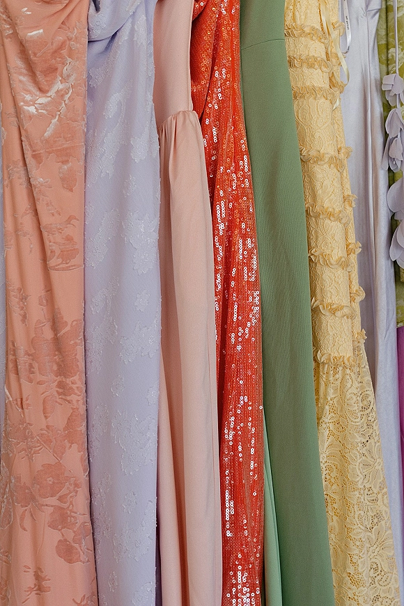 Bridesmaid dresses in mismatched pastel tones on hangers along a clothing rack, featuring lace fabric, sequins, jacquard, and ruffles
