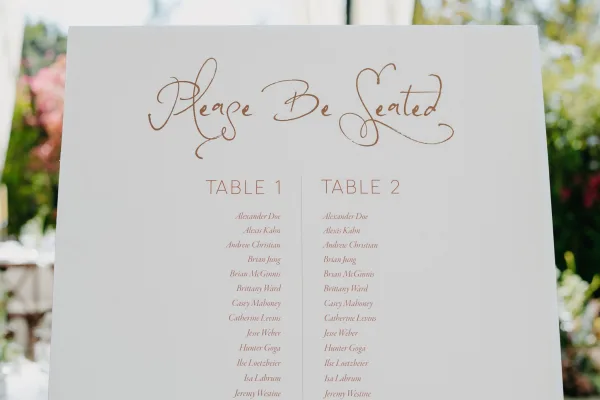Wedding seating chart sign with elegant script lettering set against garden greenery and blurred flowers in soft daylight