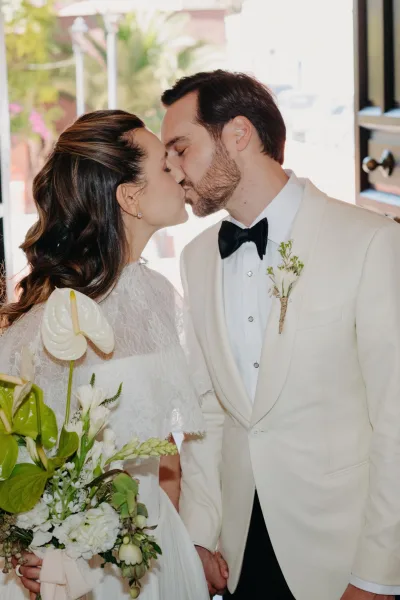 Wedding kiss portrait of a couple holding hands, the bride cradling a white bouquet, framed by a sunlit doorway with greenery and palm leaves.